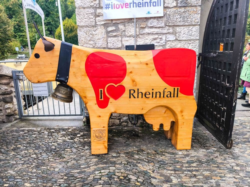 Eine große hölzerne Kuhskulptur mit einer Glocke um den Hals steht in der Nähe einer Steinmauer am Rheinfall, Europas größtem Wasserfall in Schaffhausen. Die Kuh hat rote Flecken, I ♥ Rheinfall auf der Seite, mit Fahnen und Grünzeug im Hintergrund.