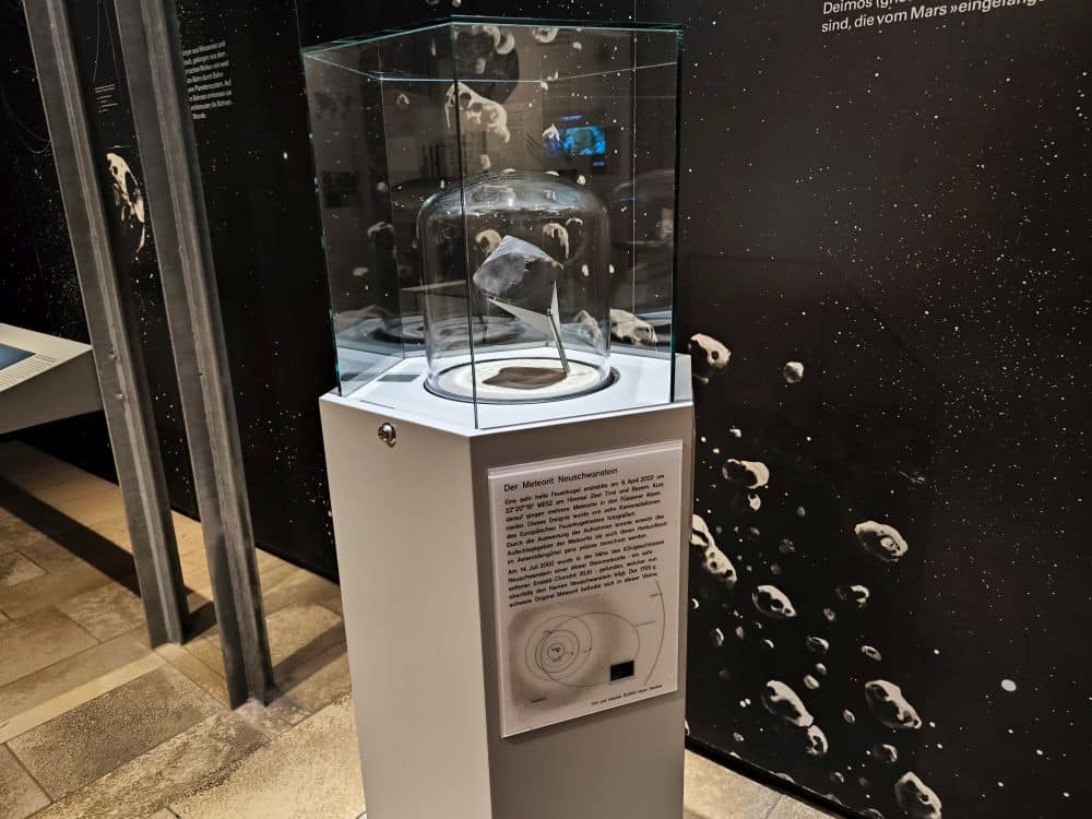 Im Rieskratermuseum in Nördlingen wird ein Meteorit in einer Glasvitrine auf einem weißen Sockel mit einer Informationstafel in deutscher Sprache ausgestellt, vor einem Hintergrund mit Bildern von Asteroiden und dem Weltraum.