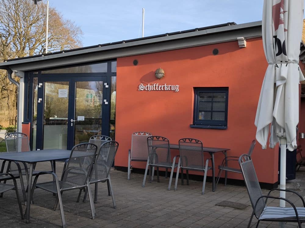 Sitzplatz im Freien mit Metalltischen und Stühlen vor einem orangefarbenen Gebäude mit der Aufschrift Schifferkrug. Genießen Sie eine entspannte Mahlzeit in einem der Lieblingsrestaurants Rügen, die mit einer Glastür, einem kleinen Fenster und einem großen weißen Terrassenschirm ausgestattet sind.