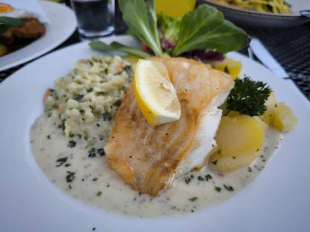 Ein Teller mit gekochtem Weißfisch, gekrönt von einer Zitronenspalte, serviert mit Sahnesoße, Kartoffelpüree, Blattgemüse und Kräutern - ein köstlicher Geschmack der Insel Rügen, den man oft in den besten Restaurants Rügens findet. Im Hintergrund sind andere Gerichte und ein Glas Wasser zu sehen.