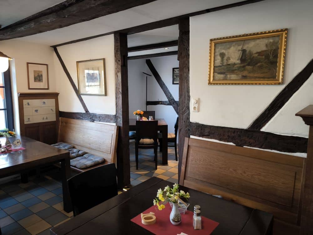 Ein gemütlicher, rustikaler Essplatz auf der Insel Rügen mit Holzbalken, Bänken und Tischen. Frische Blumen und ein rotes Tischset stehen im Vordergrund. Gemälde schmücken die weißen Wände, während warmes Tageslicht in eines der Lieblingsrestaurants Rügen fällt.