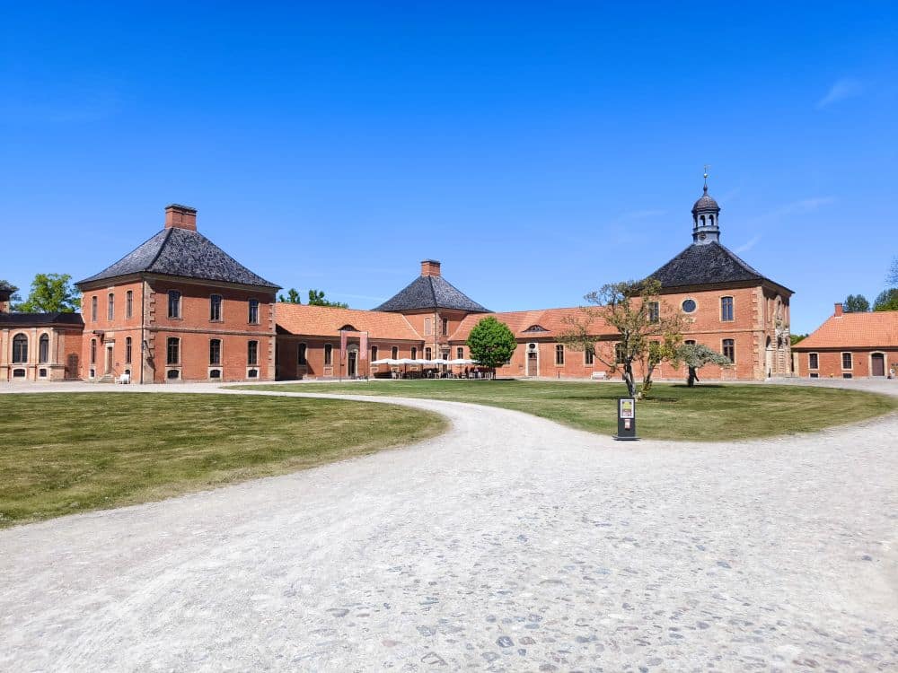 Ein großer historischer Backsteinbaukomplex in Klütz mit einem zentralen Rundturm, umgeben von grünen Rasenflächen und einem geschwungenen Kiesweg unter dem klaren blauen Himmel der Ostseeküste.