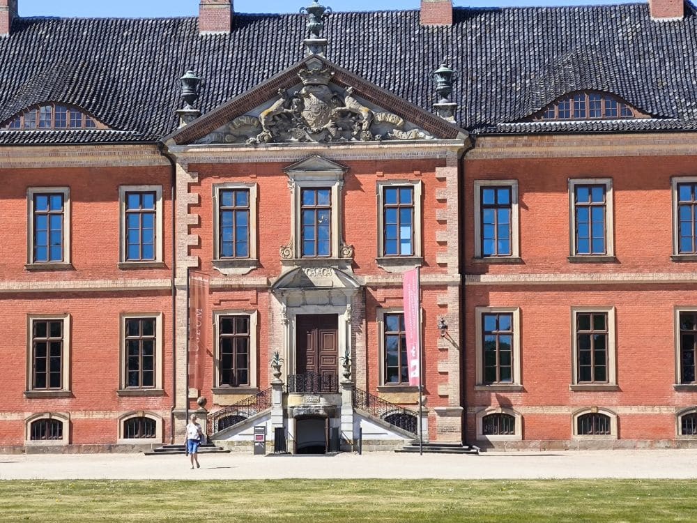 Ein großes historisches Backsteingebäude in Klütz in der Nähe der Ostseeküste mit hohen Fenstern, kunstvollen Steinverzierungen über dem Haupteingang, zwei Seitentreppen und einer Person, die auf den Stufen im hellen Sonnenlicht steht.