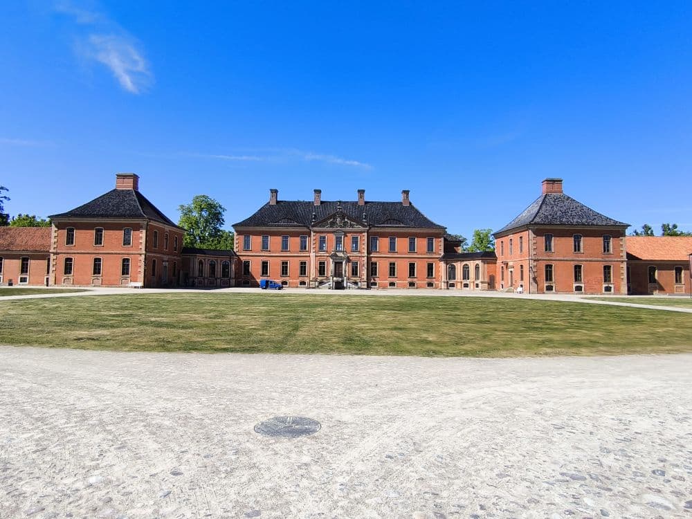 Ein großes historisches Backsteingebäude mit drei Hauptteilen und dunklen Dächern, umgeben von einem großzügigen grasbewachsenen Innenhof unter einem strahlend blauen Himmel - ein verstecktes Juwel in Klütz, perfekt, um den Charme der Ostseeküste zu entdecken.
