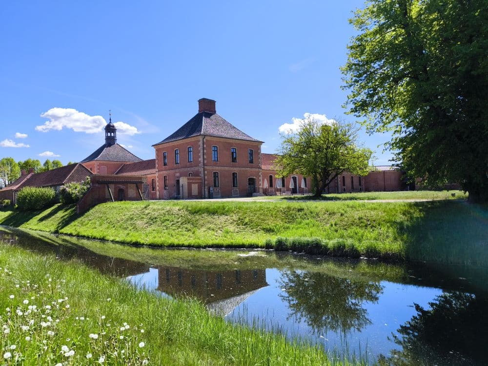 Ein großes rotes Backsteingebäude mit einem Turm steht neben einem grasbewachsenen Wassergraben in Klütz und spiegelt sich im ruhigen Wasser unter einem strahlend blauen Himmel. Üppig grüne Bäume und Gras umgeben diese friedliche Szene in der Nähe der Ostseeküste.