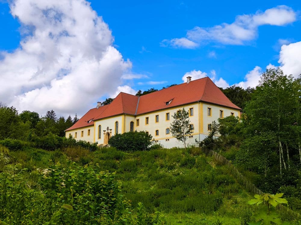 Ein großes Gebäude mit rotem Dach und cremefarbenen Wänden steht auf einem grasbewachsenen, baumbestandenen Hügel bei Hayingen unter einem strahlend blauen Himmel - ein einladender Anblick für alle, die die Hochgehschätzt Wanderung Glastal genießen.
