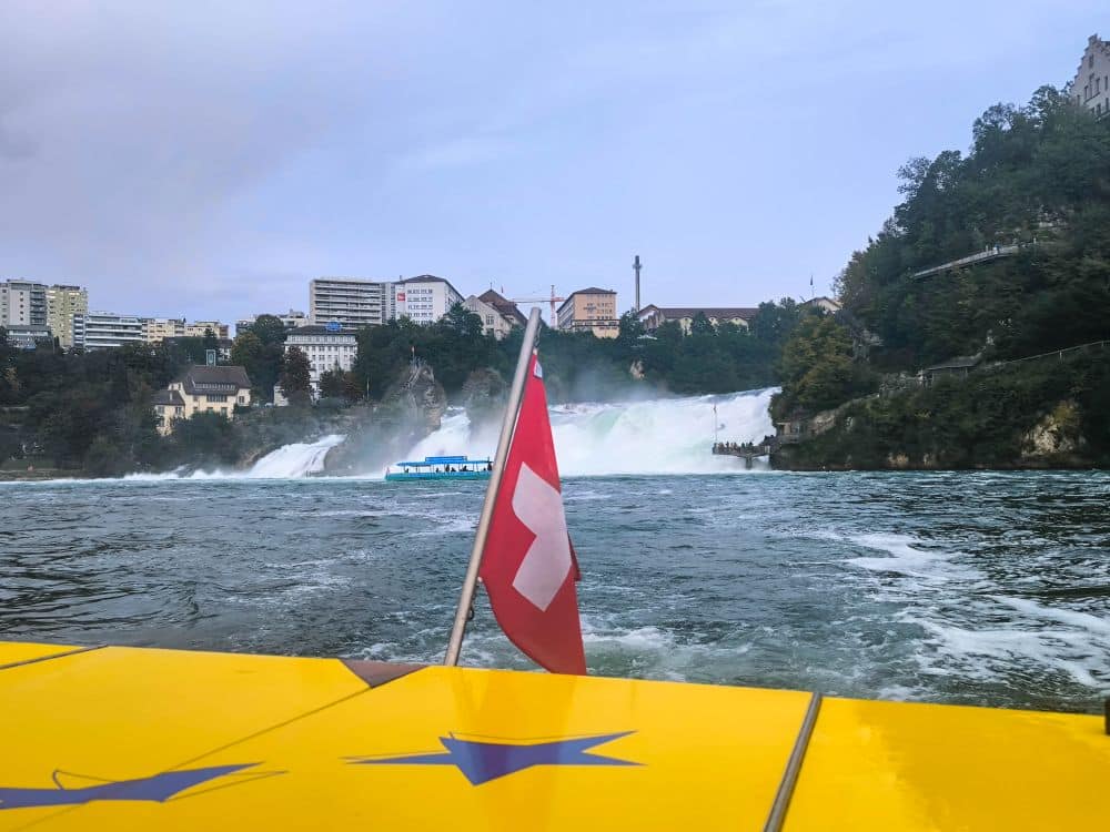Eine Schweizer Flagge auf einem gelben Boot im Vordergrund, mit dem Rheinfall - Europas größtem Wasserfall - im Hintergrund, umgeben von den Gebäuden der Stadt Schaffhausen und Grünanlagen unter einem bewölkten Himmel.