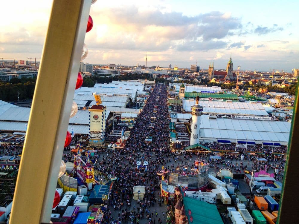 Luftaufnahme eines überfüllten Oktoberfestes mit großen weißen Zelten, belebten Gehwegen voller Menschen und verschiedenen bunten Attraktionen; im Hintergrund sind städtische Gebäude und ein teilweise bewölkter Himmel zu sehen.
