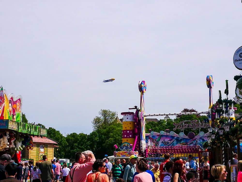 Ein belebter Rummelplatz mit bunten Ständen, Fahrgeschäften und Oktoberfestdekorationen. Viele Menschen laufen umher, und ein Luftschiff schwebt am Himmel. Bäume und blauer Himmel bilden den Hintergrund dieser lebhaften Szene. Oktoberfest