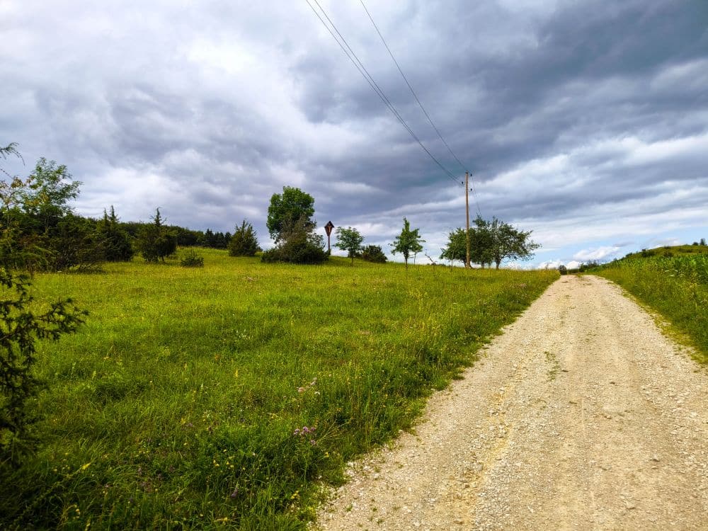 Ein Schotterweg führt unter wolkenverhangenem Himmel in der Nähe von Hayingen an einer Wiese entlang. In der Ferne stehen vereinzelte Bäume und Stromleitungen - perfekt für eine Hochgehschätzt Wanderung Glastal.