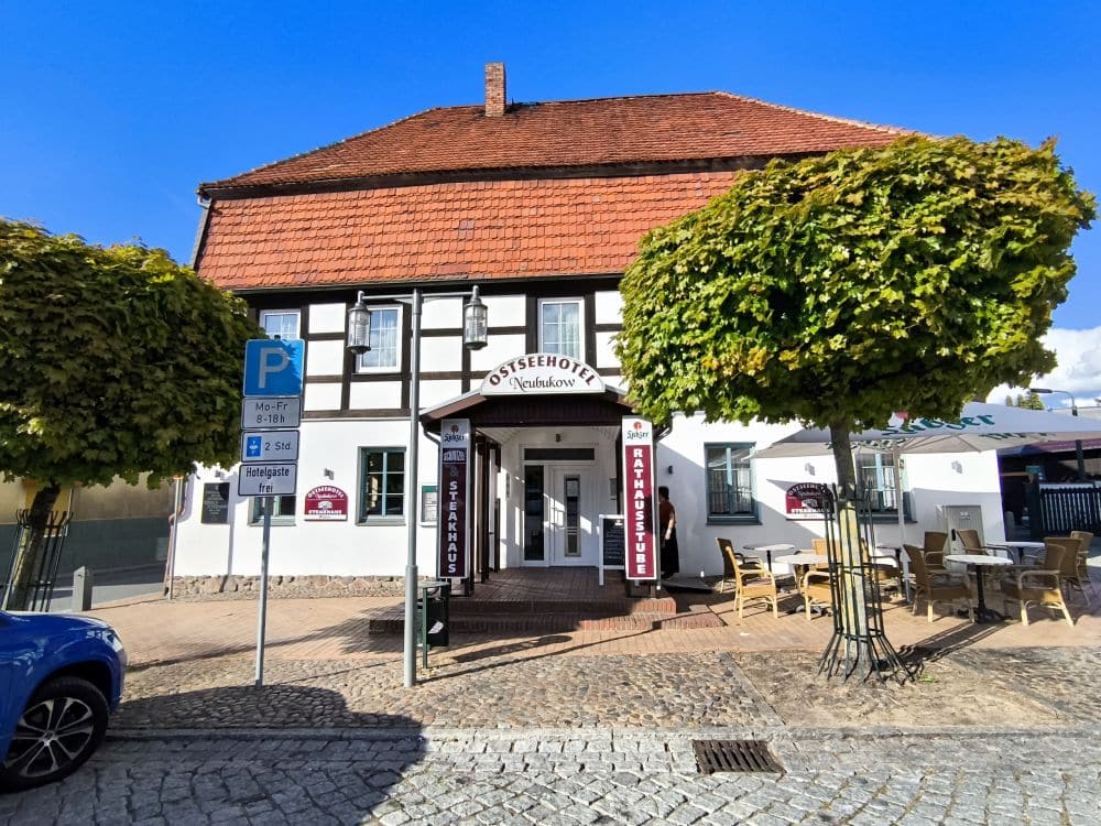 Ein traditionelles Fachwerkhaus mit rotem Ziegeldach beherbergt das Ostseehotel Neuhof in Neubukow. Tische und Stühle stehen im Freien unter beschnittenen Bäumen, und ein blaues Parkschild steht auf der Kopfsteinpflasterstraße davor.