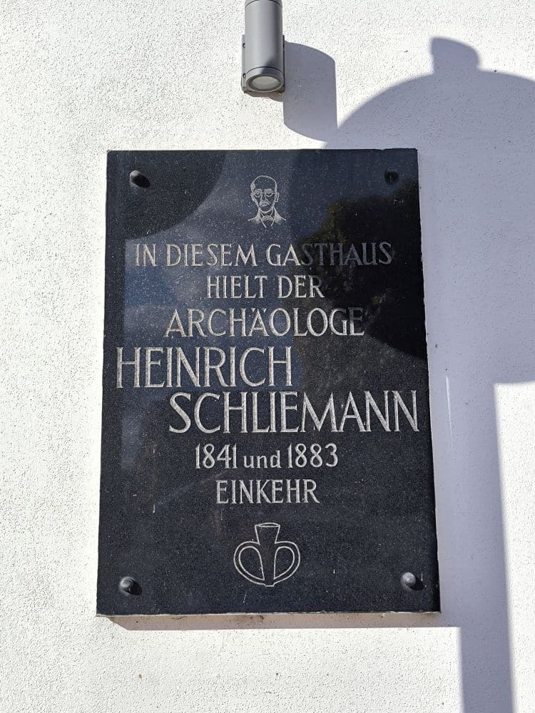 Eine schwarze Gedenktafel an einer weißen Wand in Neubukow erinnert an den Archäologen Heinrich Schliemann. Sie enthält einen Text in deutscher Sprache, seinen Namen und die Jahreszahlen (1841 und 1883) sowie eine kleine Strichzeichnung seines Gesichts über der Inschrift.