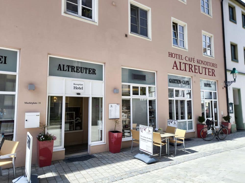 Straßenansicht des Hotels und Cafés Altreuter in Nördlingen, mit Sitzgelegenheiten im Freien, Topfpflanzen, einem Fahrrad und großen Fenstern an einem sonnigen Tag. Schilder für Hotel, Café und Konditorei sind sichtbar.