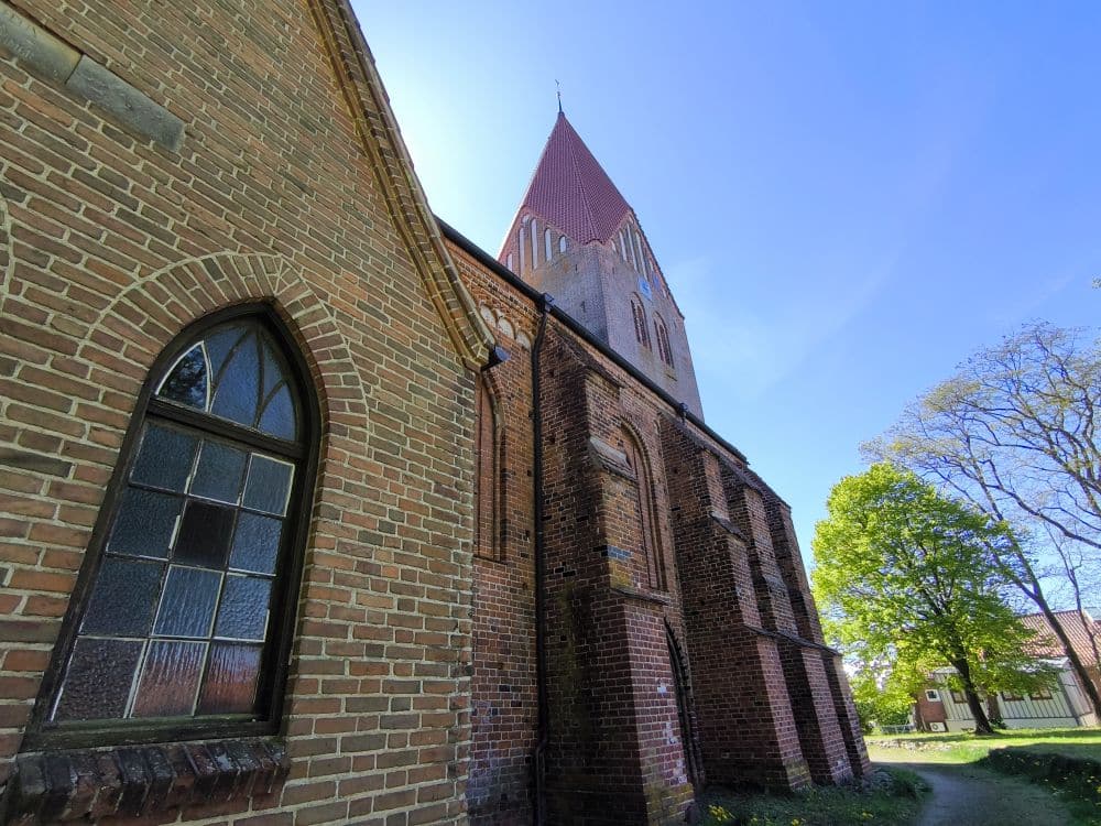 Eine Backsteinkirche mit Rundbogenfenstern und einem hohen, spitzen roten Dach steht unter einem klaren blauen Himmel in Klütz, nahe der Ostseeküste. Im Hintergrund sind grüne Bäume und ein kleines Gebäude entlang eines sonnenbeschienenen Weges zu sehen.