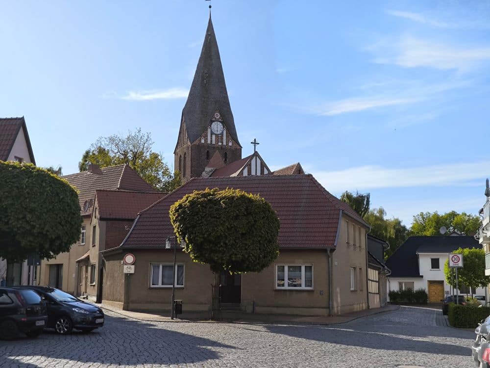 Ein kleiner Stadtplatz in Neubukow mit Kopfsteinpflaster, parkenden Autos und einer Kirche mit einem hohen Kirchturm im Hintergrund. Bäume und Häuser umgeben die Kirche, alles unter einem klaren blauen Himmel.