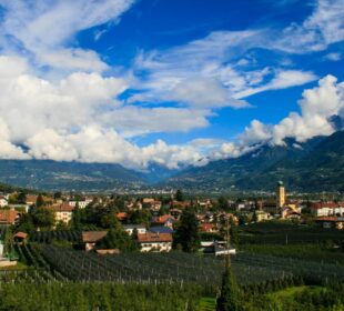 Ein malerischer Blick auf eine kleine Stadt am Brandiswaalweg, eingebettet in grüne Felder und Obstgärten, umgeben von Bergen unter einem teilweise bewölkten blauen Himmel. Rote Häuser mit Dächern und ein Kirchturm ragen aus der Landschaft heraus.
