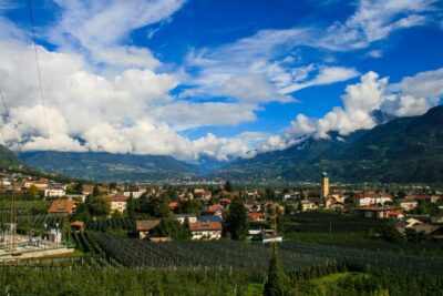 Ein malerischer Blick auf eine kleine Stadt am Brandiswaalweg, eingebettet in grüne Felder und Obstgärten, umgeben von Bergen unter einem teilweise bewölkten blauen Himmel. Rote Häuser mit Dächern und ein Kirchturm ragen aus der Landschaft heraus.