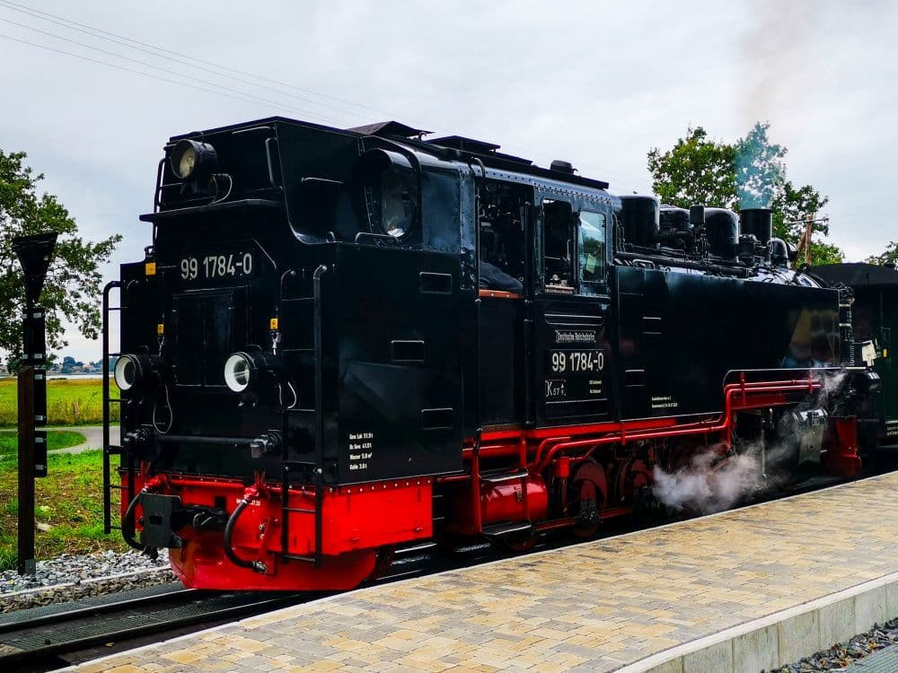 Eine schwarz-rote Dampflokomotive mit der Nummer 99 1784-0 steht auf den Gleisen einer steinernen Plattform, leichter Dampf steigt auf - das ist der ikonische Rasende Roland von Rügen, bereit zum Volldampf durch die malerischen Bäume und Felder.