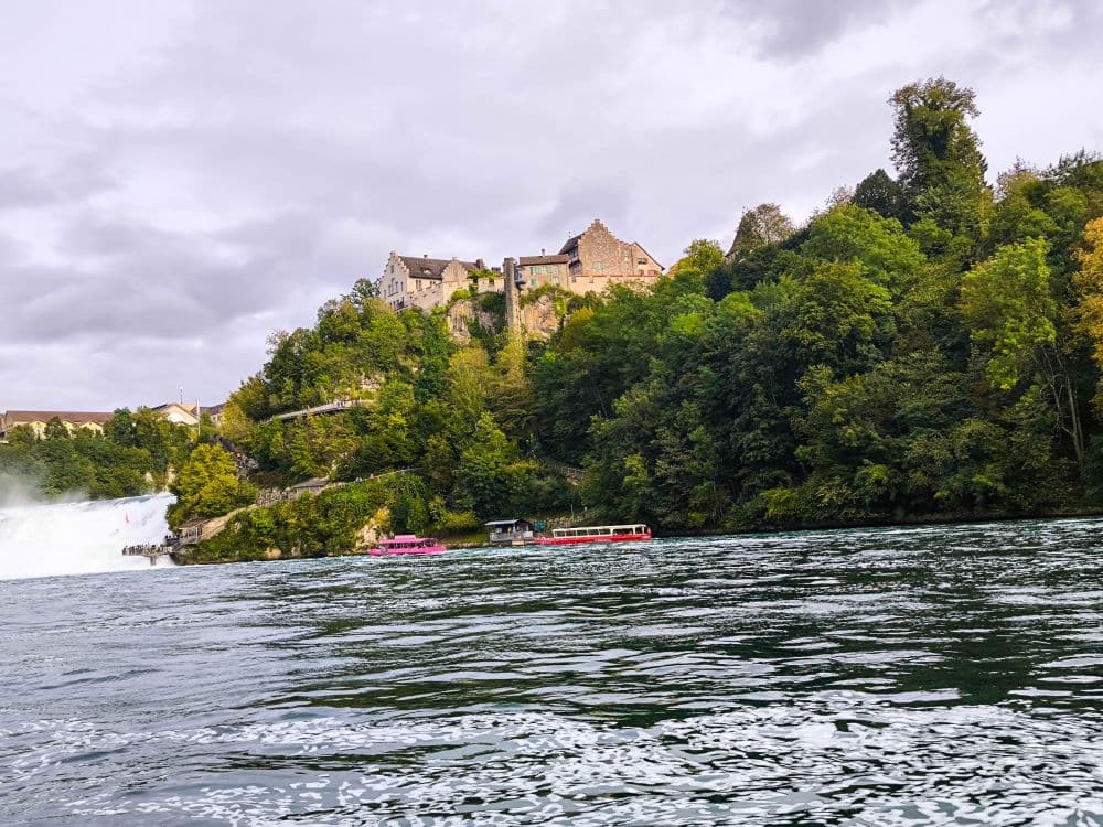 Auf einem grünen, baumbestandenen Hügel neben dem Rheinfall, dem größten Wasserfall Europas, thront ein großes Schloss über einem breiten Fluss. Zwei bunte Ausflugsboote gleiten unter dem wolkenverhangenen Schaffhauser Himmel über das Wasser.