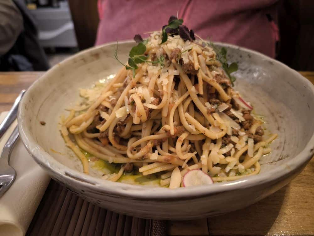 Auf einem Holztisch steht eine Schüssel mit Nudeln, die mit geriebenem Käse, Mikrogrün und Fleischstückchen belegt sind. Perfekt für den Genuss in einem der Insel Rügen Lieblingsrestaurants. Eine Gabel und eine Serviette liegen neben der Schüssel, während der Hintergrund sanft verschwimmt.