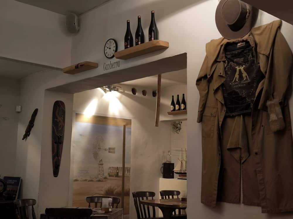 Ein gemütliches Café-Interieur auf der Insel Rügen mit Holzstühlen und -tischen, Vintage-Dekor, Weinflaschen in Regalen und einer Wandmalerei mit Windmühle und Wüste. Die warme Beleuchtung trägt zur einladenden Atmosphäre eines der Lieblingsrestaurants Rügen bei.