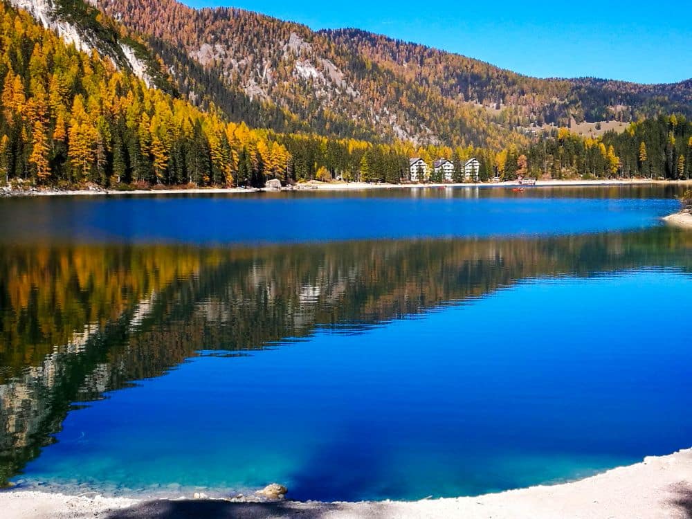 Ein klarer blauer See, bekannt als Pragser Wildsee, spiegelt die bunten Herbstbäume und Berge wider, und am anderen Ufer sind einige Gebäude unter einem strahlend blauen Himmel zu sehen.