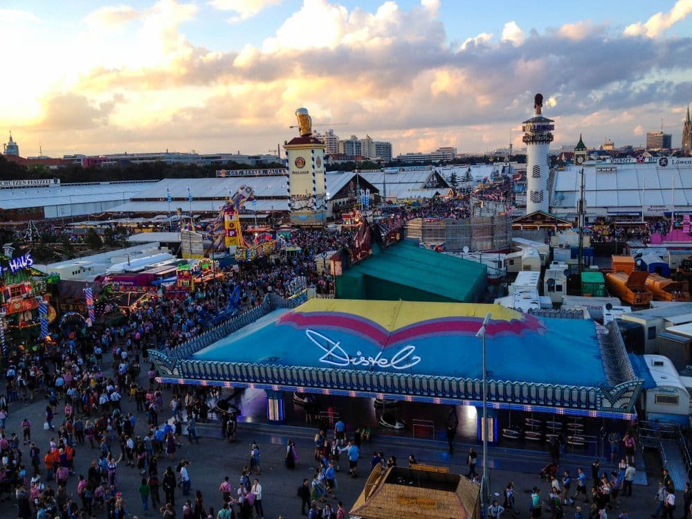Eine große Menschenmenge versammelt sich auf einem Oktoberfest im Freien mit bunten Zelten, Fahrgeschäften und Ständen unter einem teilweise bewölkten Himmel bei Sonnenuntergang. Ein blaues Zelt mit einem Dachl darauf ist im Vordergrund zu sehen.