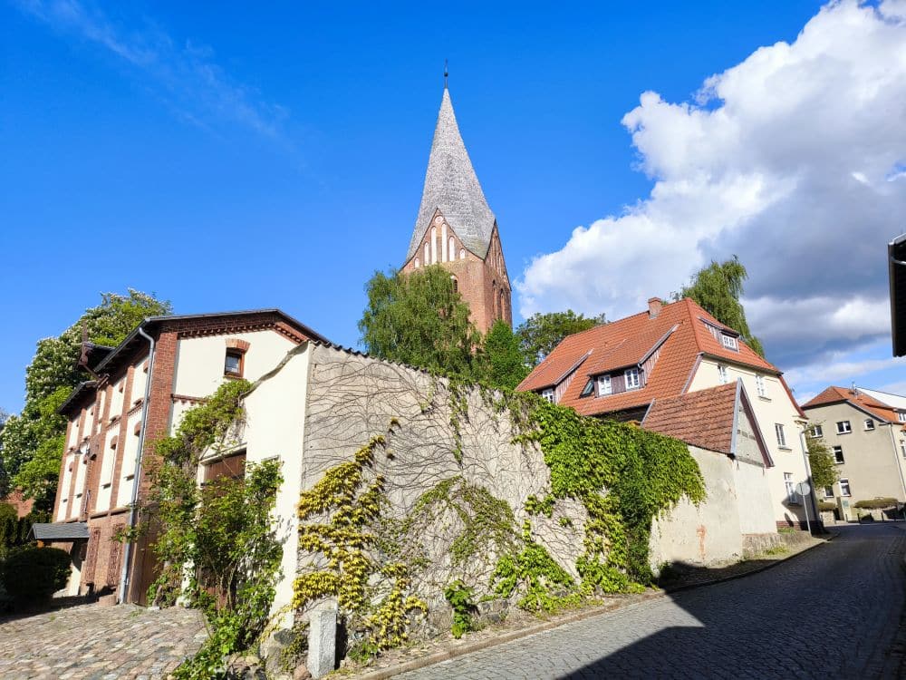 Eine von Häusern und einer efeubewachsenen Mauer gesäumte Kopfsteinpflasterstraße schlängelt sich durch Neubukow; im Hintergrund erhebt sich ein hoher, spitzer Kirchturm vor einem strahlend blauen Himmel mit einigen Wolken.