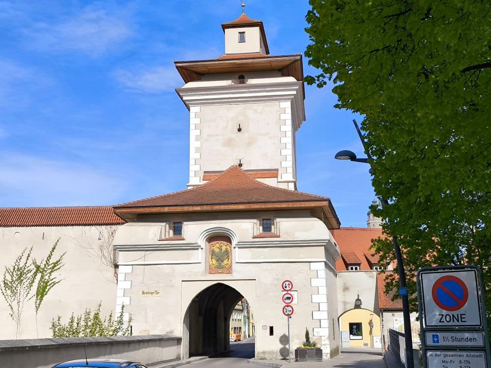 Ein historischer Torturm aus weißem Stein mit rotem Ziegeldach in Nördlingen steht unter einem klaren blauen Himmel, eingerahmt von einem grünen Laubbaum. In der Nähe der gewölbten Einfahrt sind Straßenschilder und ein Parkverbotsschild zu sehen.