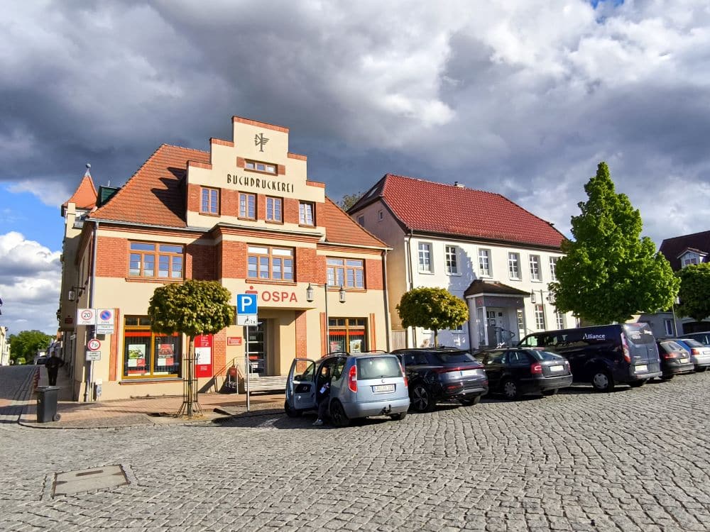 Auf einer Kopfsteinpflasterstraße in Neubukow parken Autos vor einem historischen Gebäude mit rotem Dach, das mit Buchdruckerei und OSPA beschriftet ist. Bäume säumen den Gehweg, während Wolken über dem blauen Himmel schweben.