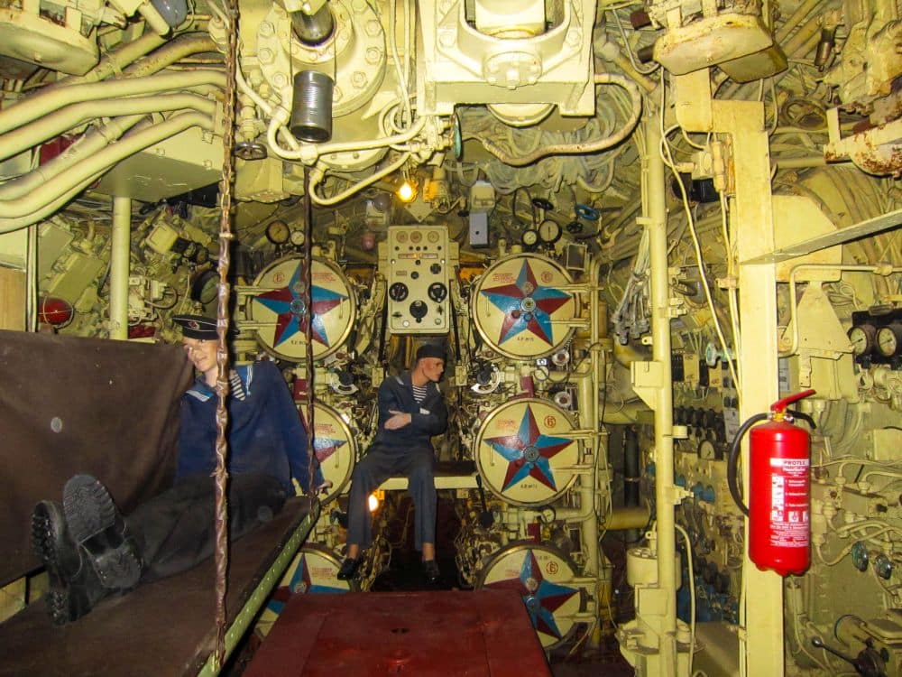 Zwei als Matrosen verkleidete Schaufensterpuppen sitzen in einem überfüllten gelben U-Boot-Kontrollraum, der an Peenemünde erinnert und voller Rohre, Zifferblätter, Schalttafeln und eines roten Feuerlöschers an der rechten Wand ist.