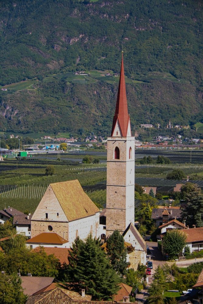 Am Brandiswaalweg steht eine steinerne Kirche mit einem hohen, roten Kirchturm zwischen Bäumen und Dächern, vor einer Kulisse aus grünen Feldern und bewaldeten Bergen.