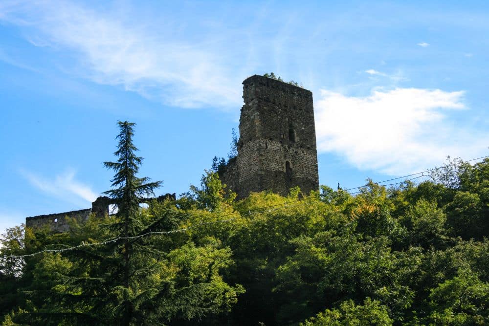 Ein Steinturm einer alten Burg am Brandiswaalweg erhebt sich über dichte grüne Bäume unter einem strahlend blauen Himmel mit Wolkenfetzen. Das Bauwerk wirkt verwittert und teilweise ruiniert.