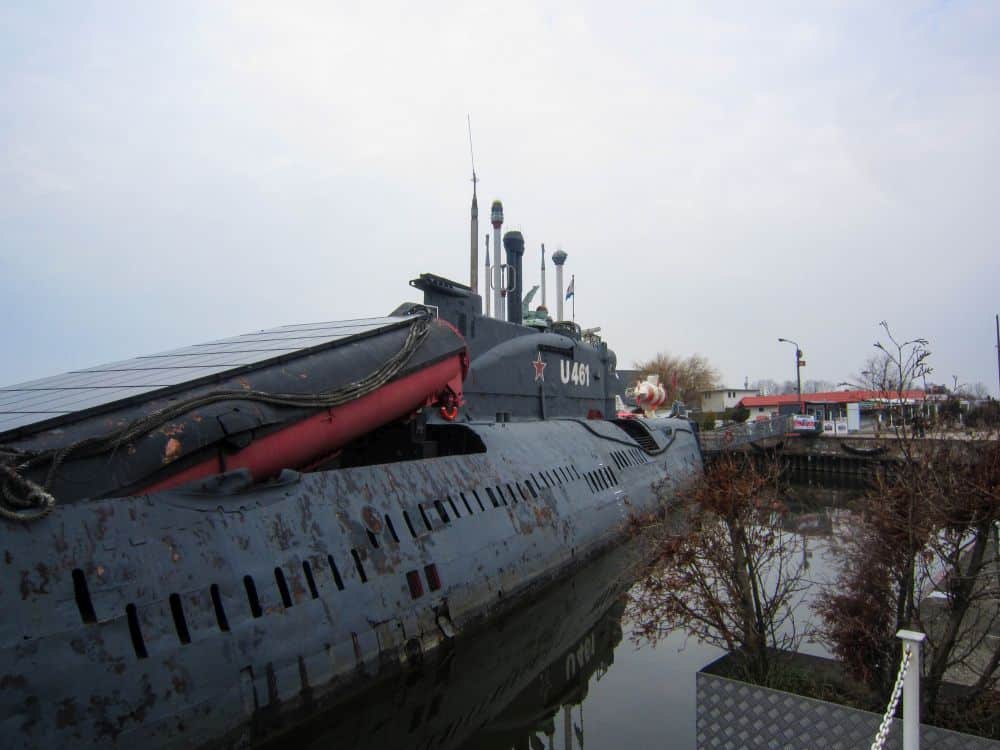Ein großes, dunkelgraues U-Boot mit der Kennzeichnung U461 liegt in der Nähe von Peenemünde an der Küste, umgeben von Bäumen und Gebäuden, und auf seinem Deck sind bei bewölktem Himmel Antennentürme und Geräte zu sehen.