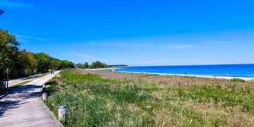 Eine hölzerne Promenade am Ostseeküsten-Radweg führt entlang der grasbewachsenen Dünen in Boltenhagen zu einem Sandstrand mit ein paar Menschen in der Ferne, gesäumt von der leuchtend blauen Ostsee und einem klaren, wolkenlosen Himmel - wahrlich ein Paradies am Meer.