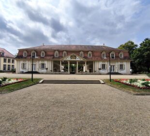 Ein prächtiges historisches Gebäude mit säulenbestücktem Eingang, symmetrischem Aufbau und rotem Ziegeldach steht umgeben von gepflegten Gärten, Blumenbeeten und Natur und zeigt historische Schätze unter einem wolkenverhangenen Himmel.