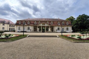 Ein prächtiges historisches Gebäude mit säulenbestücktem Eingang, symmetrischem Aufbau und rotem Ziegeldach steht umgeben von gepflegten Gärten, Blumenbeeten und Natur und zeigt historische Schätze unter einem wolkenverhangenen Himmel.