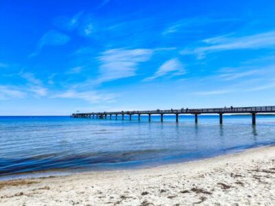 Eine lange Holzmole erstreckt sich über das ruhige, klare blaue Wasser des Ostseeparadieses in Boltenhagen unter einem strahlend blauen Himmel mit Wolkenfetzen. Im Vordergrund ist ein weicher Sandstrand mit Algenflecken zu sehen.