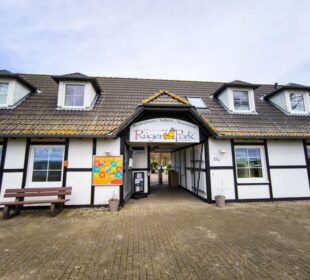 Eingang zu einem weißen Gebäude mit dunkler Holzverkleidung und einem Schild mit der Aufschrift "Rügenpark" über der Eingangstür. Draußen stehen Gaubenfenster, eine braune Bank und eine bunte Pinnwand unter dem klaren Himmel.