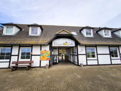 Eingang zu einem weißen Gebäude mit dunkler Holzverkleidung und einem Schild mit der Aufschrift "Rügenpark" über der Eingangstür. Draußen stehen Gaubenfenster, eine braune Bank und eine bunte Pinnwand unter dem klaren Himmel.