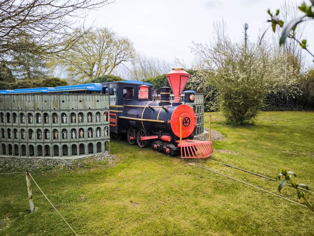 Eine kleine, bunte Spielzeugeisenbahn mit einer rot-schwarzen Lokomotive fährt auf schmalen Schienen durch eine grüne Wiese im Rügenpark, die teilweise von einem niedrigen, geschwungenen Zaun umgeben ist. Bäume und Sträucher umgeben die Szene.