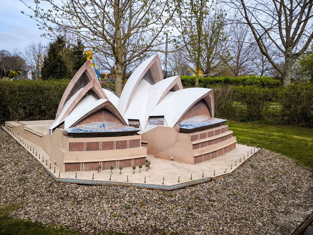 Ein kleines Außenmodell des Sydney Opera House im Rügenpark, umgeben von Kies und Gras, mit Bäumen und Sträuchern im Hintergrund. Das Modell bildet die ikonische weiße segelartige Dachkonstruktion des Gebäudes nach.