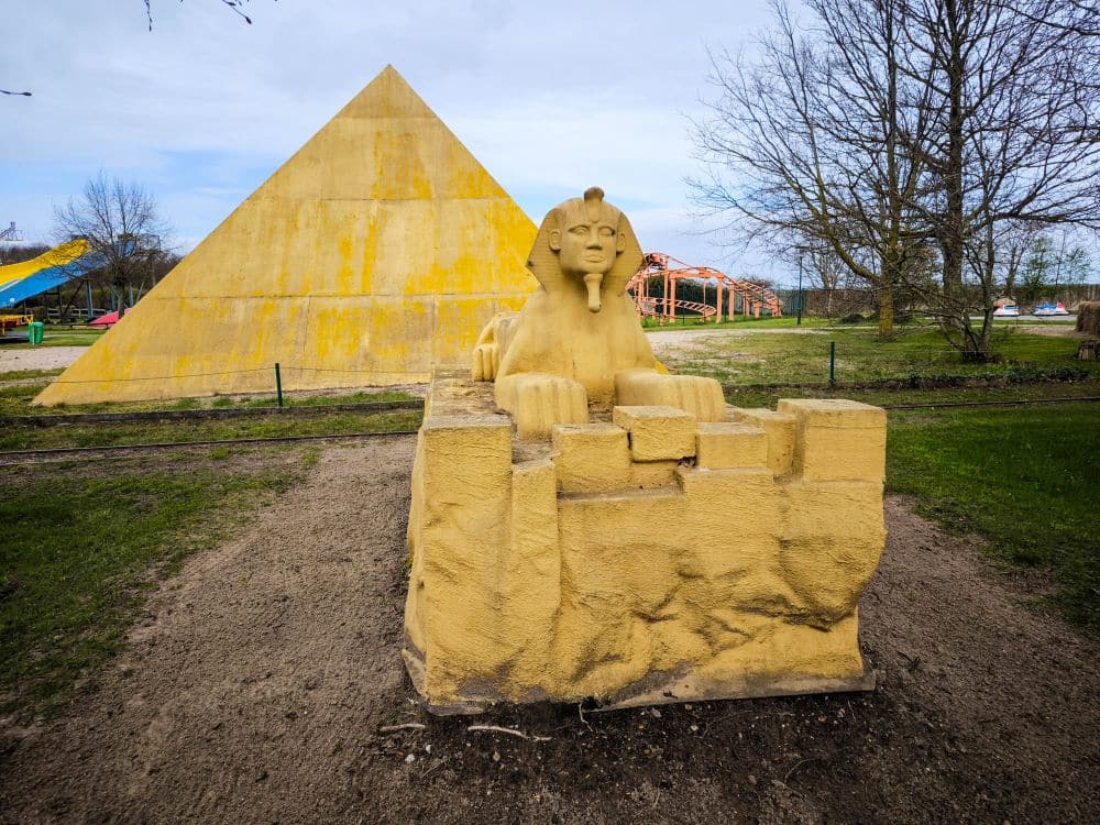 Eine gelbe Steinnachbildung der Sphinx steht vor einem großen Pyramidenbau im Rügenpark, eingebettet in einen grasbewachsenen Park mit Bäumen und Fahrgeschäften im Hintergrund.