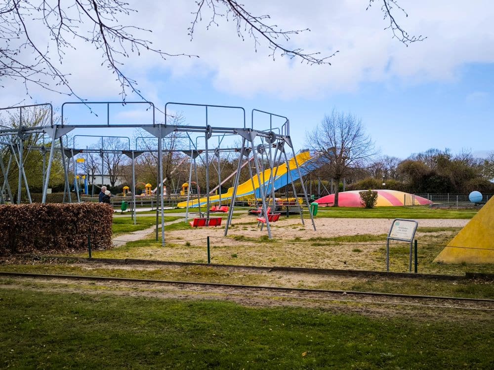Auf dem Spielplatz im Rügenpark gibt es ein großes Metallklettergerüst, Wippen, eine gelbe Rutsche und einen bunten, kuppelförmigen Spielbereich auf Sand, alles umgeben von Gras, Bäumen und Zäunen unter einem teilweise bewölkten Himmel.