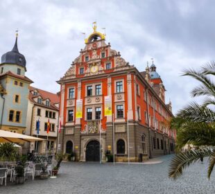 Ein leuchtend rotes und beiges historisches Gebäude mit verschnörkelten Details steht an einer Kopfsteinpflasterstraße in Gotha, flankiert von anderen alten Gebäuden. Unter dem wolkenverhangenen Himmel sind Sitzgelegenheiten im Freien und Palmen zu sehen.