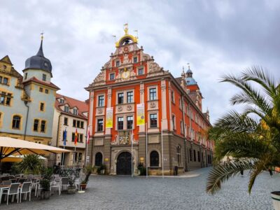 Ein leuchtend rotes und beiges historisches Gebäude mit verschnörkelten Details steht an einer Kopfsteinpflasterstraße in Gotha, flankiert von anderen alten Gebäuden. Unter dem wolkenverhangenen Himmel sind Sitzgelegenheiten im Freien und Palmen zu sehen.