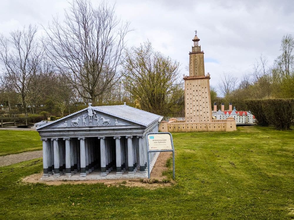 Miniaturmodelle eines antiken griechischen Tempels und eines hohen Leuchtturms stehen im Freien auf der Wiese des Rügenparks, mit Bäumen und bewölktem Himmel im Hintergrund. Vor dem Modell des Tempels steht ein kleines Schild.