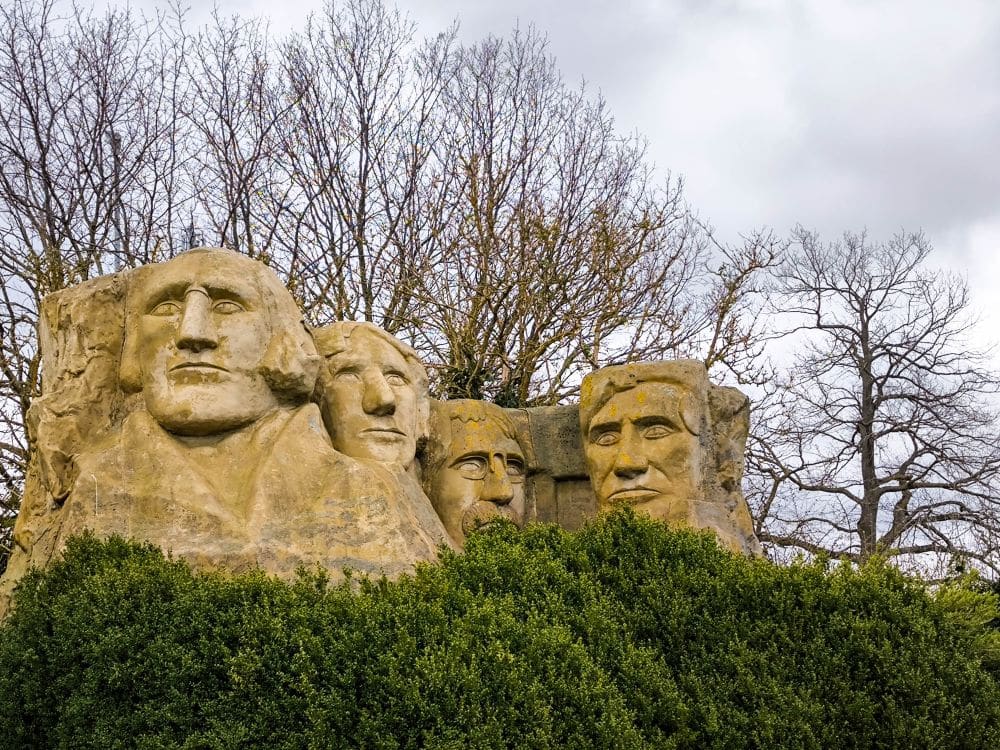 Vier große Steinbüsten, die an Präsidenten erinnern, sind teilweise hinter grünen Büschen im Rügenpark versteckt. Laublose Bäume und ein wolkenverhangener Himmel verstärken die Mount Rushmore-ähnliche Atmosphäre.