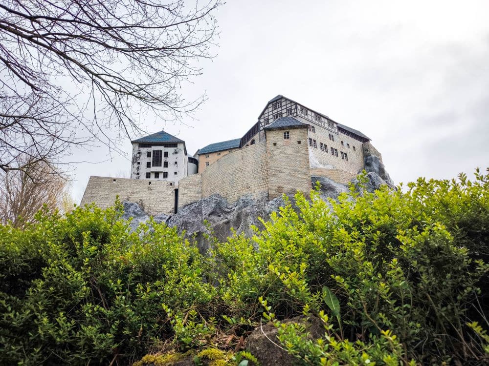 Ein großes Steinschloss mit mehreren Dächern thront auf einem felsigen Hügel im Rügenpark, umgeben von grünen Büschen und Bäumen unter einem bewölkten Himmel.