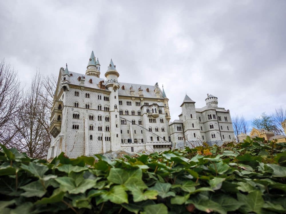 Ein prächtiges weißes Steinschloss mit mehreren Türmen und Spitzen, das an die Miniaturen des Rügenparks erinnert, steht unter einem wolkenverhangenen Himmel, der aus einem niedrigen Winkel hinter grünem, efeubewachsenem Boden zu sehen ist. Laublose Bäume rahmen die Szene ein.